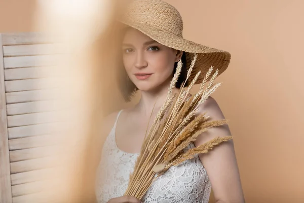 Mujer en sombrero de paja y vestido sosteniendo espiguillas cerca de la pantalla plegable aislado en beige - foto de stock