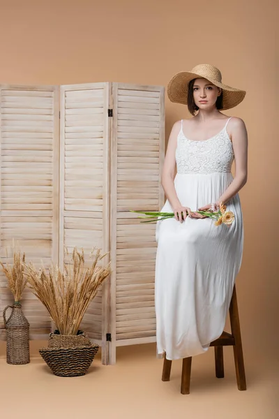 Mujer embarazada en sombrero de sol sosteniendo flores mientras está sentada cerca de la pantalla plegable en beige - foto de stock