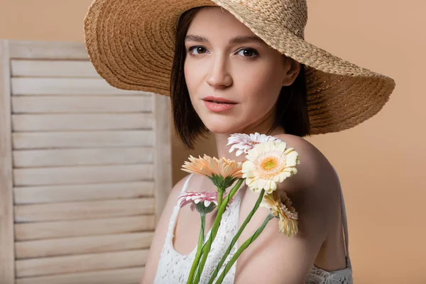 Portrait de femme au chapeau de soleil tenant des fleurs près d'un écran pliant flou isolé sur beige — Photo de stock