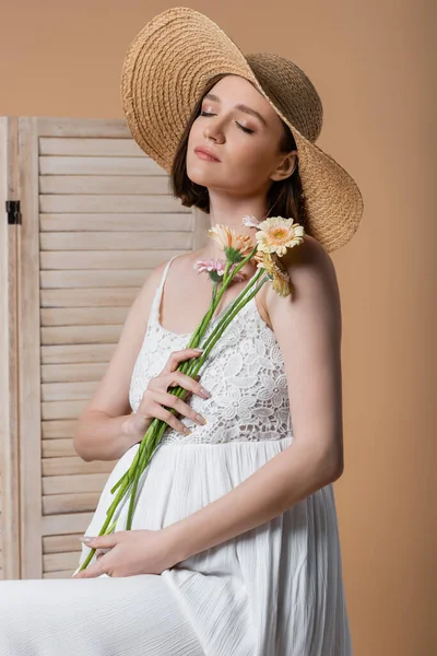 Mulher grávida em chapéu de sol segurando flores perto de tela dobrável isolado no bege — Fotografia de Stock