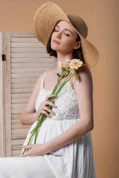Pregnant woman in sun hat holding flowers near folding screen isolated on beige — Stock Photo