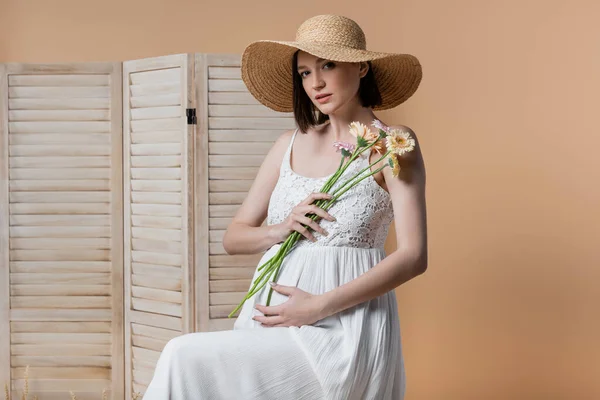 Donna incinta in abito che tiene fiori vicino schermo pieghevole isolato su beige — Foto stock