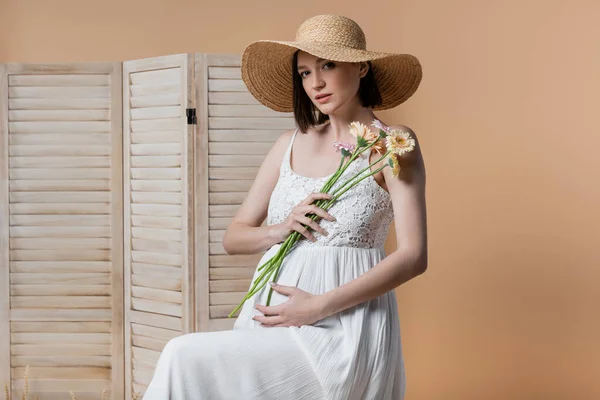 Pregnant woman in dress holding flowers near folding screen isolated on beige — Stock Photo