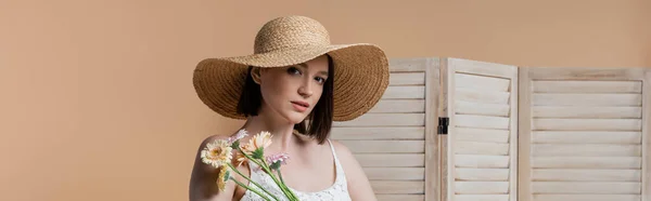 Mujer morena en sombrero de paja mirando a la cámara cerca de flores y pantalla plegable aislada en beige, pancarta - foto de stock