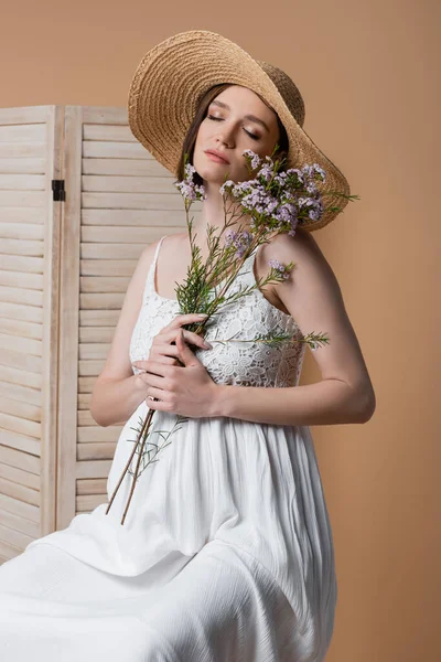 Mujer embarazada en sombrero de paja cerrando los ojos y sosteniendo la planta cerca de la pantalla plegable aislada en beige - foto de stock