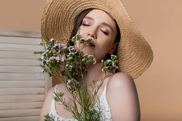 Frau mit Strohhut schließt Augen und hält Pflanzen isoliert auf Beige — Stockfoto