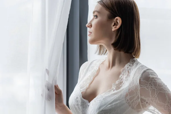 Вид сбоку женщины в кружевном халате, стоящей у окна дома — стоковое фото