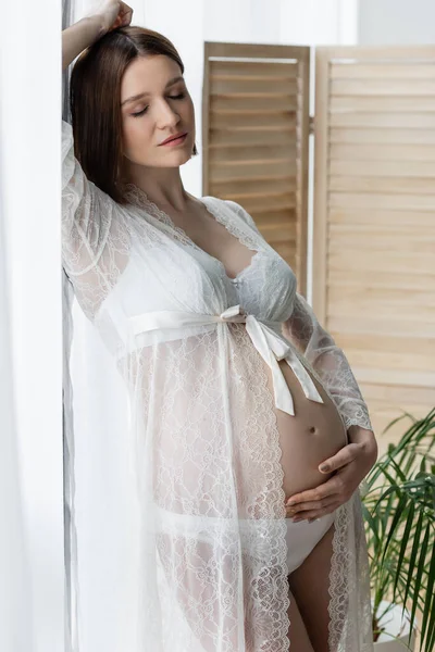 Mujer embarazada en ropa interior y túnica de pie cerca de las cortinas en casa - foto de stock