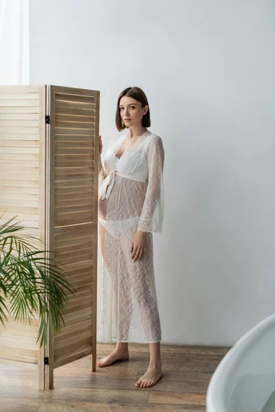 Barefoot pregnant woman in robe standing near folding screen in bathroom — Stock Photo