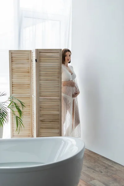 Pregnant woman in robe looking at camera near folding screen and bathtub with water — Stock Photo