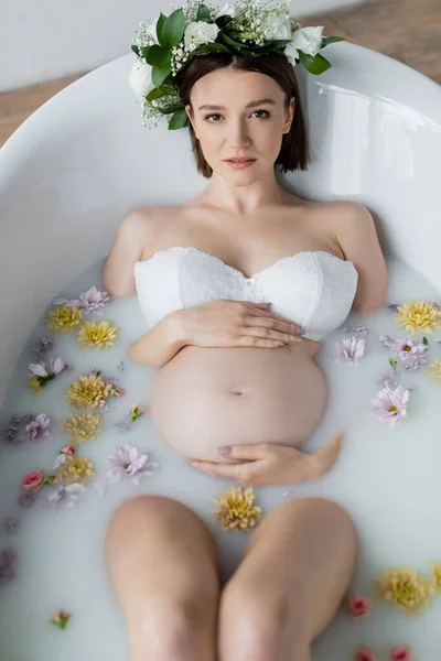 Vista dall'alto di donna incinta in ghirlanda che si trova in vasca da bagno con fiori e latte — Foto stock