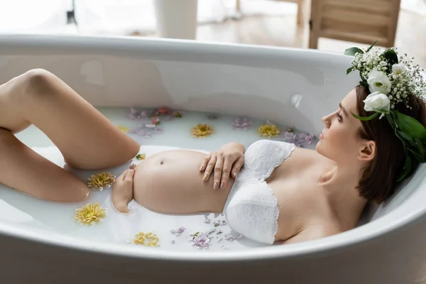 Vista laterale della donna incinta in ghirlanda sdraiata in bagno con fiori e latte a casa — Foto stock
