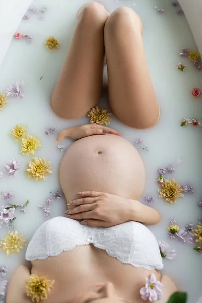 Vista superior de la mujer embarazada tocando el vientre en el baño con flores y leche - foto de stock
