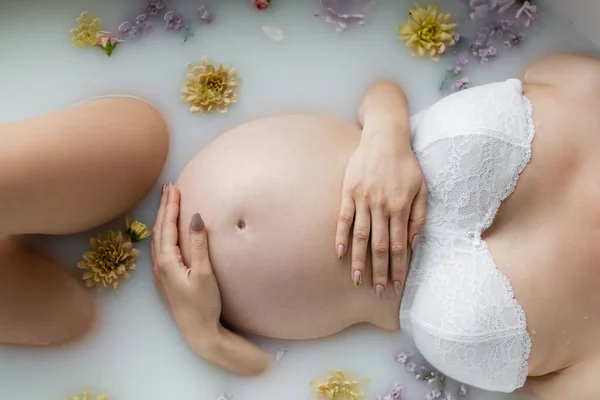 Cropped view of pregnant woman in bra lying in milk bath with plants at home — Stock Photo