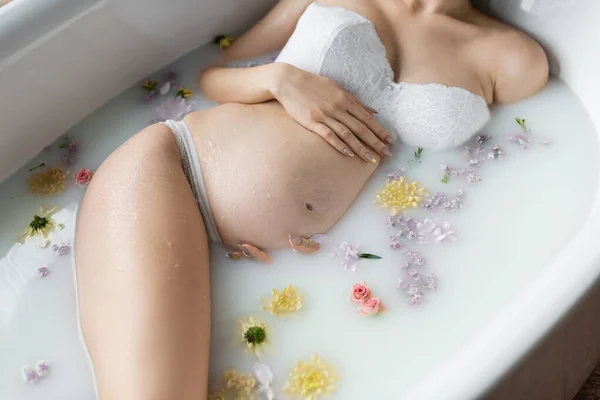 Vista recortada de la mujer embarazada en lencería tocando el vientre en baño con flores y leche — Stock Photo