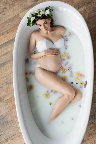 Вид сверху на беременную женщину в цветочном венке, принимающую молочную ванну дома — стоковое фото