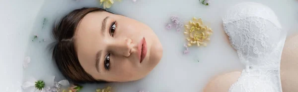 Vista superior de la mujer joven en sujetador mirando a la cámara en baño de leche con flores, pancarta - foto de stock