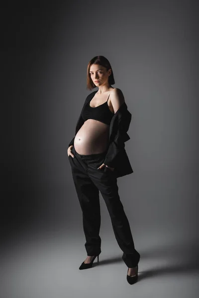 Longitud completa de la mujer embarazada con estilo en tacones y traje de pie sobre fondo gris — Stock Photo