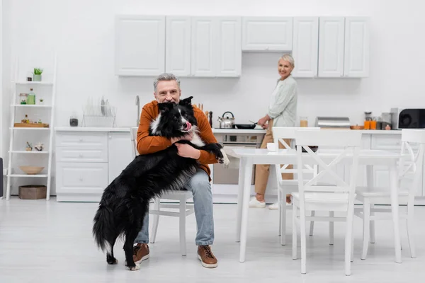 Mature man petting border collie near blurred wife in kitchen — Stock Photo