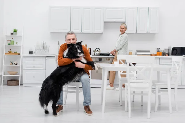 Sourire femme âgée cuisine et regarder mari avec collie frontière dans la cuisine — Photo de stock