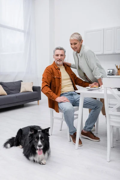 Cheerful couple looking at border collie near breakfast in kitchen — Stock Photo