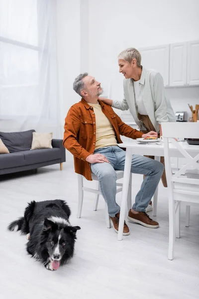 Smiling couple looking at each other near breakfast and border collie in kitchen — Stock Photo