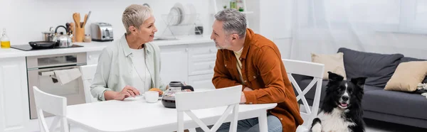 Смолящая пожилая женщина разговаривает с мужем за кофе, завтраком и пограничным колли на кухне, баннер — стоковое фото