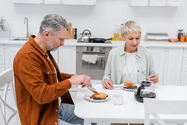 Uomo maturo taglio frittelle vicino moglie e caffè in cucina — Foto stock