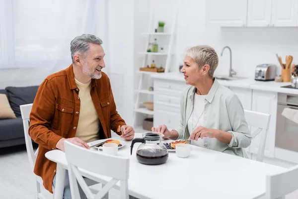 Cheerful mature man talking to wife near tasty pancakes and coffee in kitchen — Stock Photo