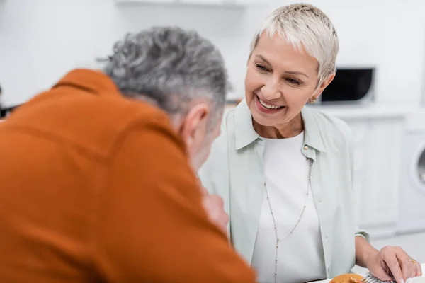 Alegre mujer mayor mirando borrosa marido cerca de panqueques en la cocina - foto de stock