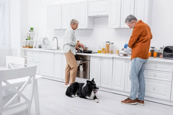 Vue latérale de l'homme versant de la nourriture pour chien dans un bol près de la frontière collie et femme cuisinant dans la cuisine — Photo de stock