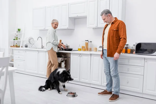 Sorrindo casal olhando para fronteira collie sentado perto de tigela com comida na cozinha — Fotografia de Stock