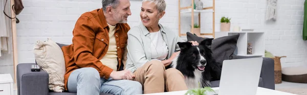 Smiling couple talking and petting border collie near laptop at home, banner — Stock Photo
