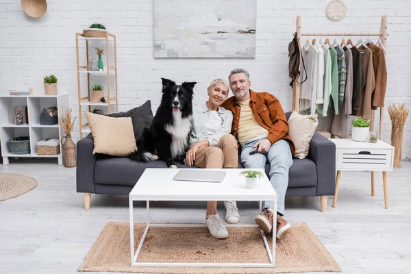 Couple positif et frontière collie chien regardant la caméra près de l'ordinateur portable sur la table basse à la maison — Photo de stock