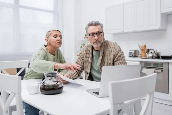 Ältere Frau zeigt auf Laptop neben Ehemann mit Papier und Kaffee in Küche — Stockfoto