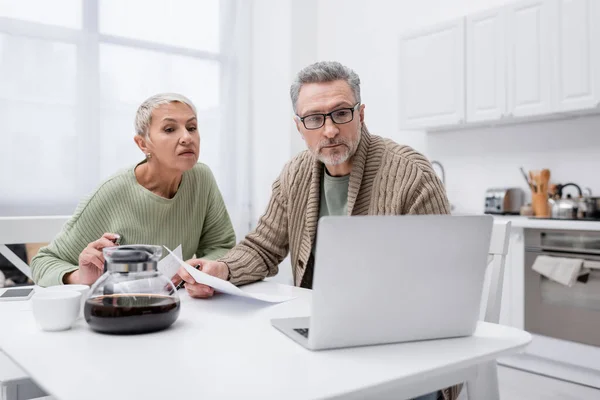 Rentner blickt auf Laptop und hält Papiere neben Kaffee in der Küche — Stockfoto