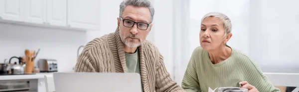 Mature man looking at blurred laptop near wife in kitchen, banner — Stock Photo