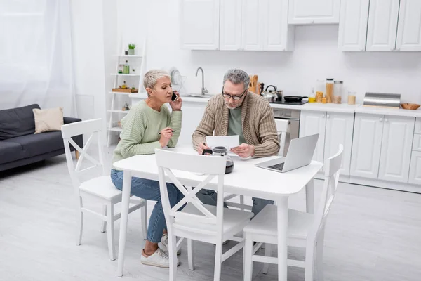 Donna anziana che parla su smartphone mentre il marito tiene la carta in cucina — Foto stock