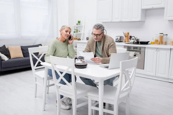 Donna anziana che parla su smartphone mentre il marito tiene la carta vicino a caffè e laptop in cucina — Foto stock