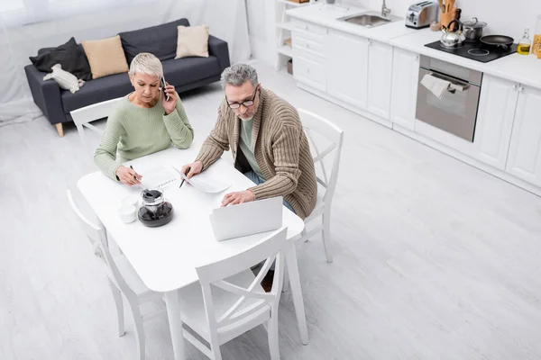 Вид сверху на зрелого мужчину, держащего бумагу и использующего ноутбук, пока жена разговаривает по мобильному телефону на кухне — стоковое фото