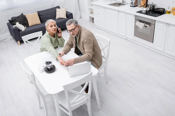 Vista aérea de la mujer mayor hablando por teléfono celular y apuntando a la computadora portátil cerca del marido con papel en la cocina - foto de stock