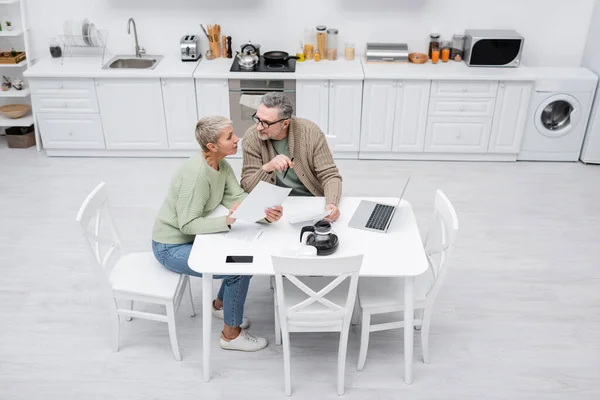 Visão de alto ângulo de pensionistas segurando papéis e falando perto de café e gadgets na cozinha — Fotografia de Stock