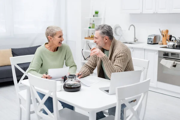 Веселая пожилая пара разговаривает рядом с бумагами и ноутбуком на кухне — стоковое фото