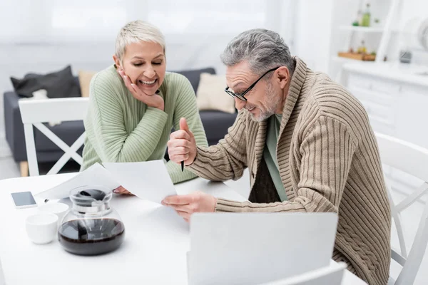 Lächelnder Rentner hält Dokument in der Nähe seiner Frau, Kaffee und Geräte in der Küche — Stockfoto