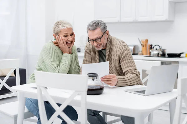 Fröhliche Seniorin schaut in Küche neben Ehemann und Laptop auf Dokument — Stockfoto