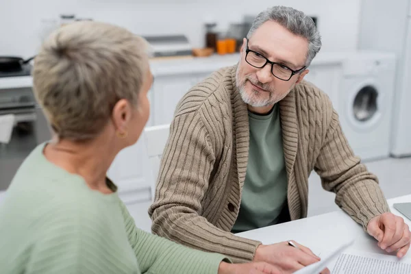 Reifer Mann sieht verschwommene Frau in der Nähe von Dokumenten in Küche an — Stockfoto