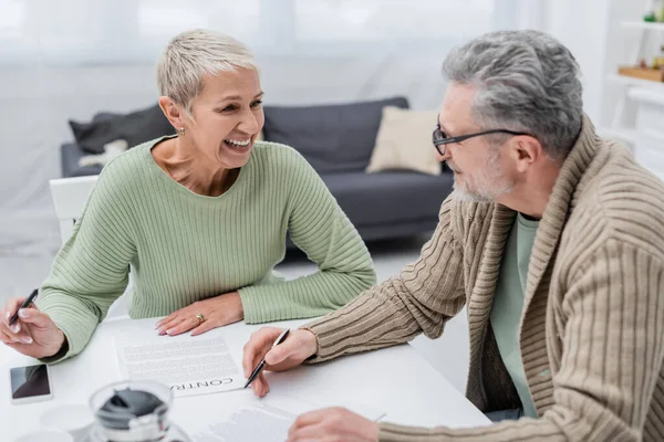 Positive senior woman looking at husband near contract and smartphone in kitchen — Stock Photo