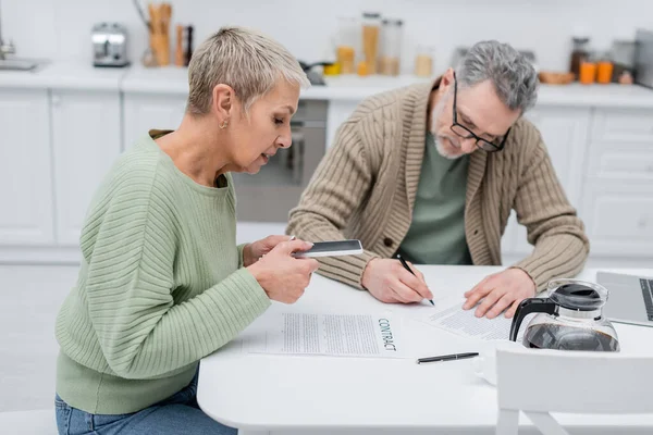 Senior woman taking photo of contract near husband and coffee in kitchen — Stock Photo