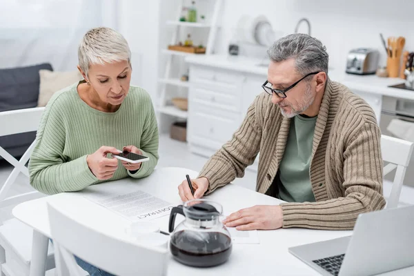 Pensionista escribiendo en el documento cerca de la esposa tomando la foto del contrato en la cocina - foto de stock