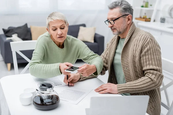 Senior woman taking photo of document near husband and coffee in kitchen — Stock Photo